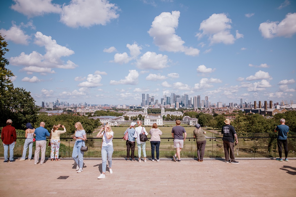 A view from the General Wofle statue looking down toward the Queen's House. White clouds pepper a blue sky and people stand with their back to the camera as they look down at the view