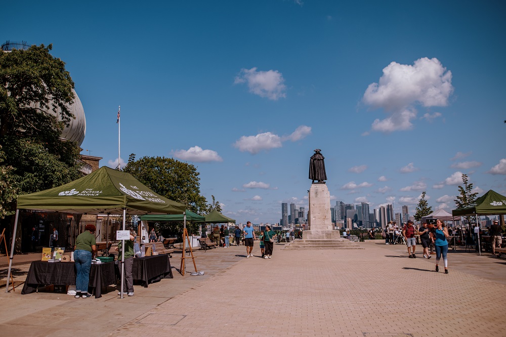 A view of the new piazza by the General Wolfe statue. The statue is on the right and a Royal Parks gazebo on the left, people walk and sit on the square in the sunshine.