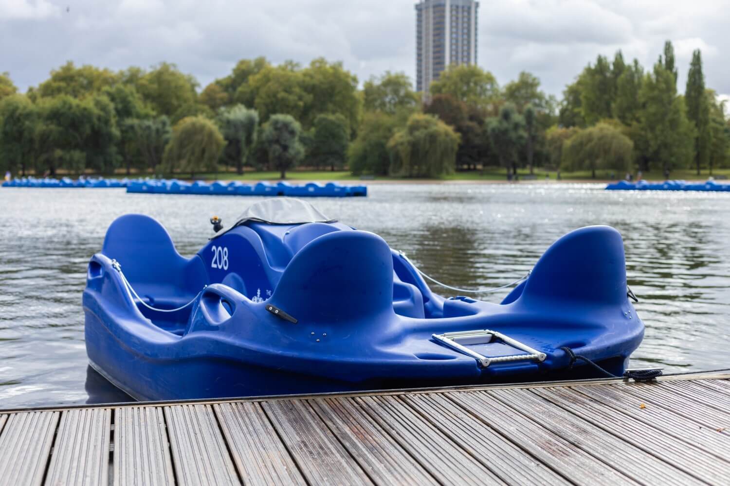 A single electric pedalo sits on the water by the wooden jetty on the Serpentine Lake in Hyde Park. Trees are visible in the background. 