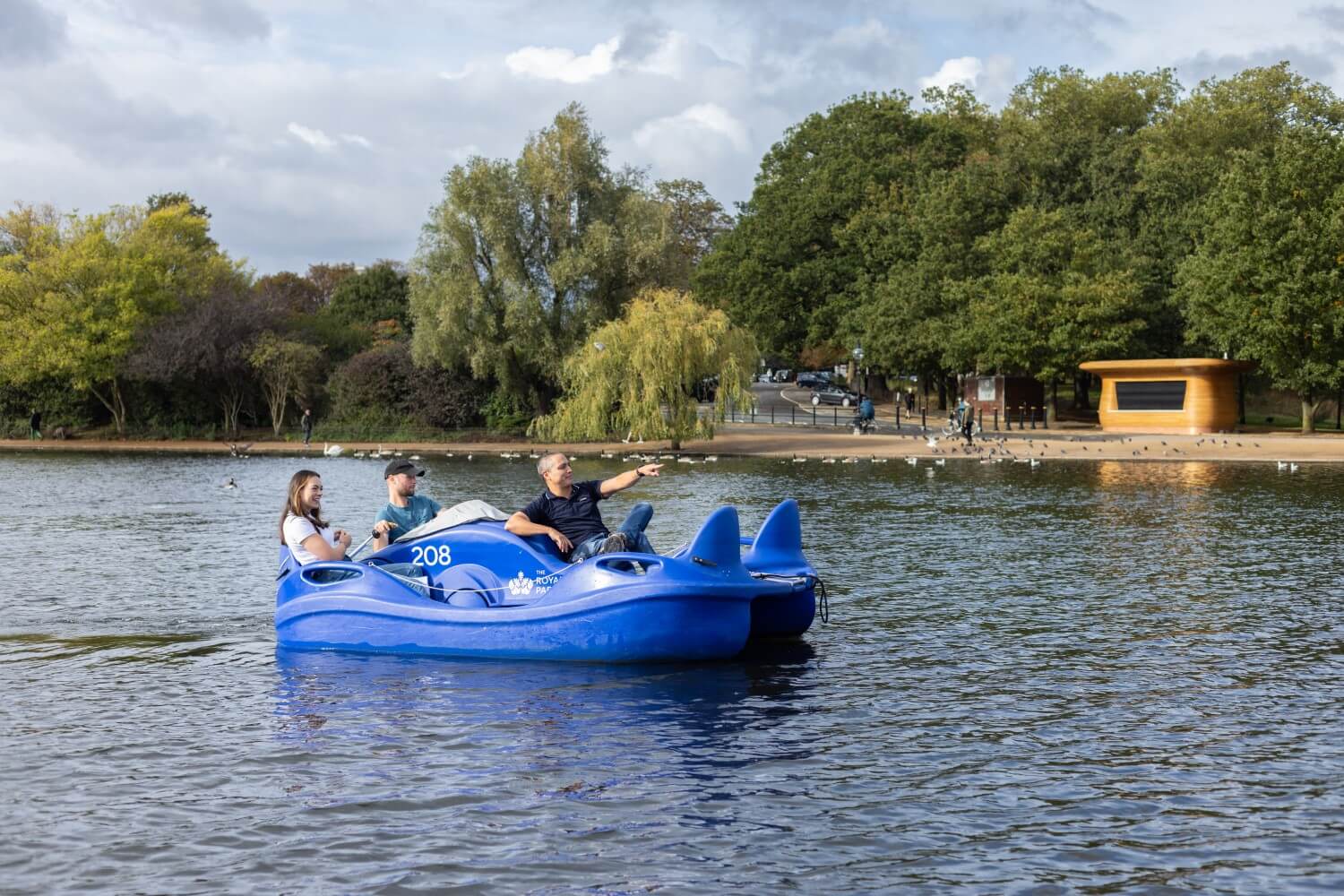 Three people ride an electric pedalo in Hyde Park on the Serpentine lake. There is a kiosk and trees in the background. The person at the front of the pedalo is pointing to something in the distance.  