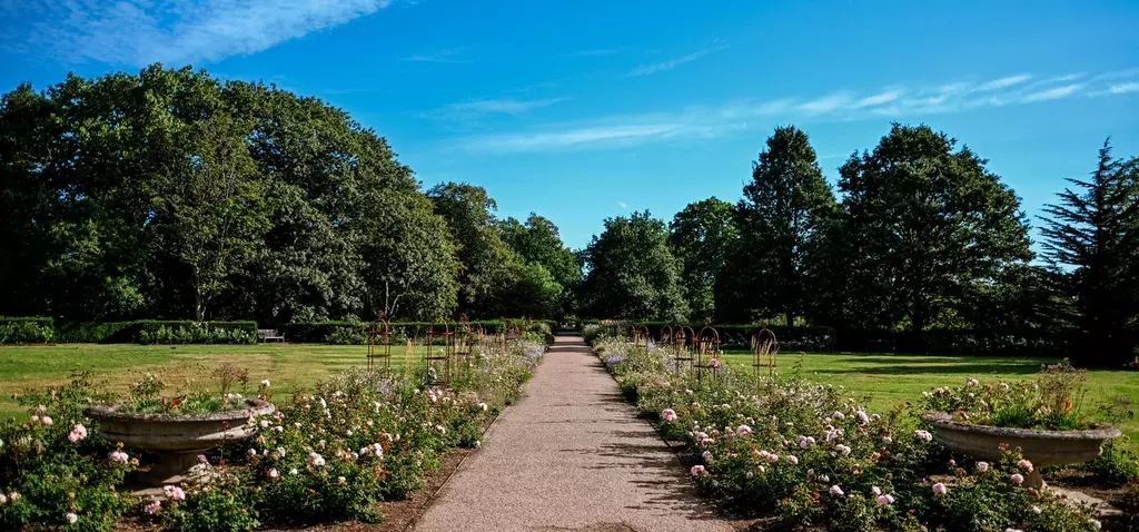 A wide pathway runs through a garden with flower beds of pink and white blossoms on either side. Stone planters and green lawns surround the path, while tall trees and a bright blue sky with light clouds. 