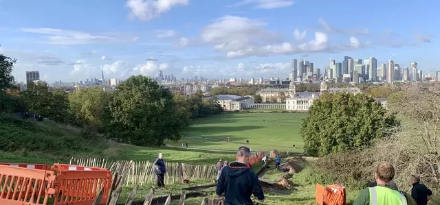 A view looking down the Grand Ascent over Greenwich Park toward Canary Wharf, it is a sunny day and the sky is blue as the volunteers start digging the trench in the foreground.