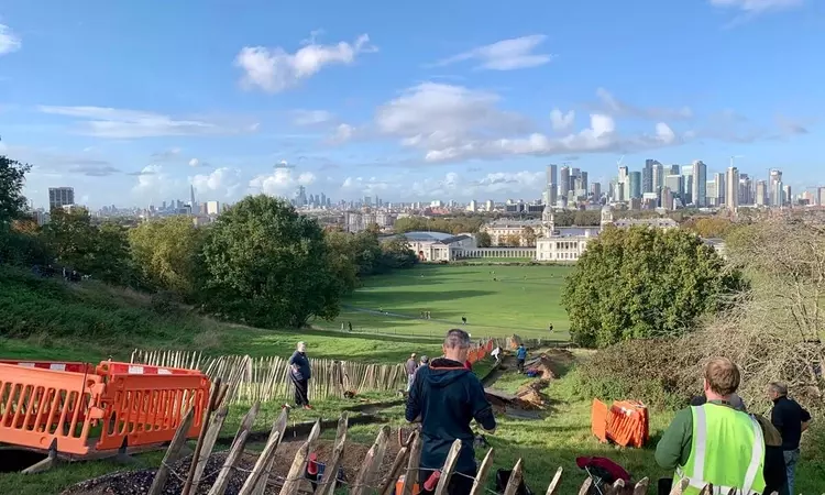 A view looking down the Grand Ascent over Greenwich Park toward Canary Wharf, it is a sunny day and the sky is blue as the volunteers start digging the trench in the foreground.