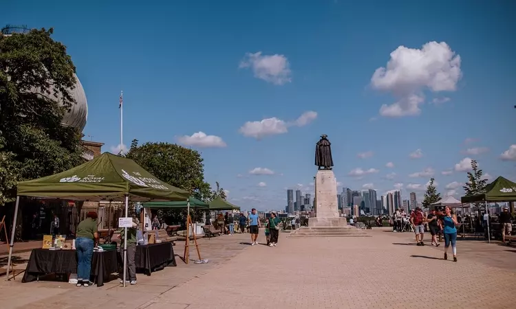 A view of the new piazza by the General Wolfe statue. The statue is on the right and a Royal Parks gazebo on the left, people walk and sit on the square in the sunshine.