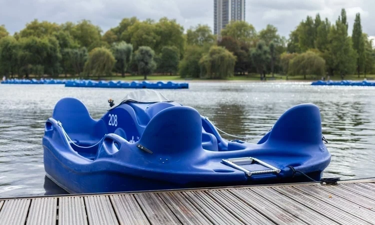A single electric pedalo sits on the water by the wooden jetty on the Serpentine Lake in Hyde Park. Trees are visible in the background.