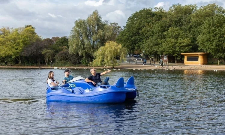 Three people ride an electric pedalo in Hyde Park on the Serpentine lake. There is a kiosk and trees in the background. The person at the front of the pedalo is pointing to something in the distance.