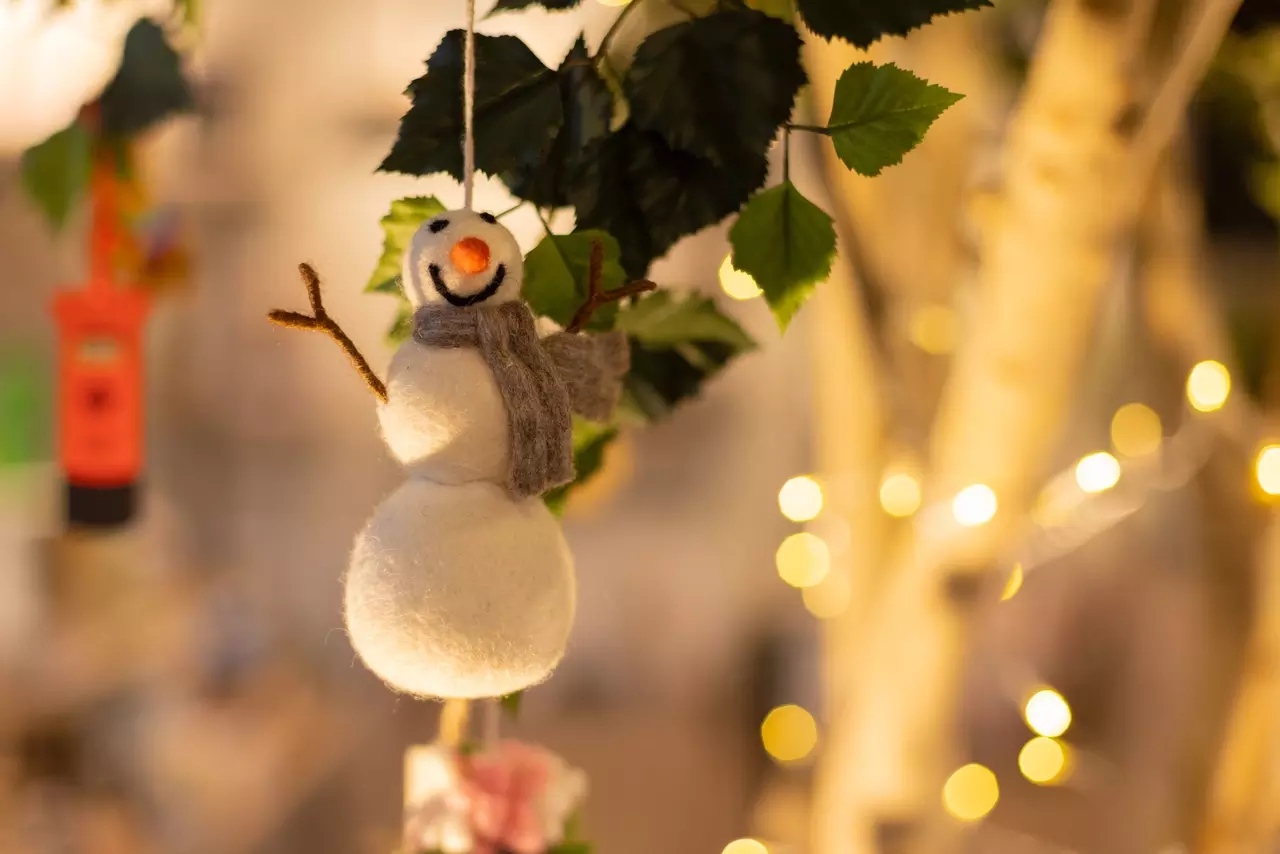 A snowman tree decoration made from felt hangs with fairy lights in the background