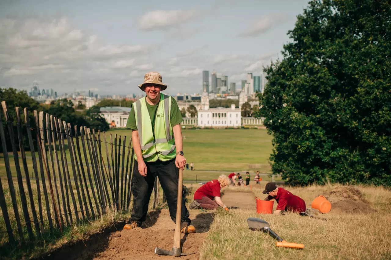Community archaeologist Andrew Mayfield stands on the Grand Ascent with the Queen's House and Canary Wharf behind him as he holds a spade and digs the trench.