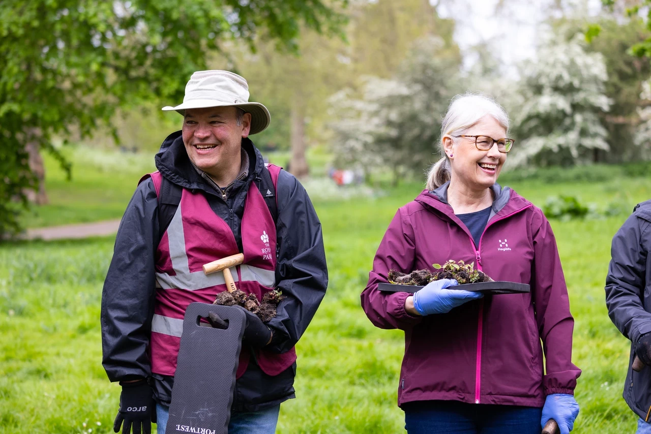 Two people smiling, looking in opposite directions. They are wearing burgundy vests and carrying various tools required for planting bulbs.