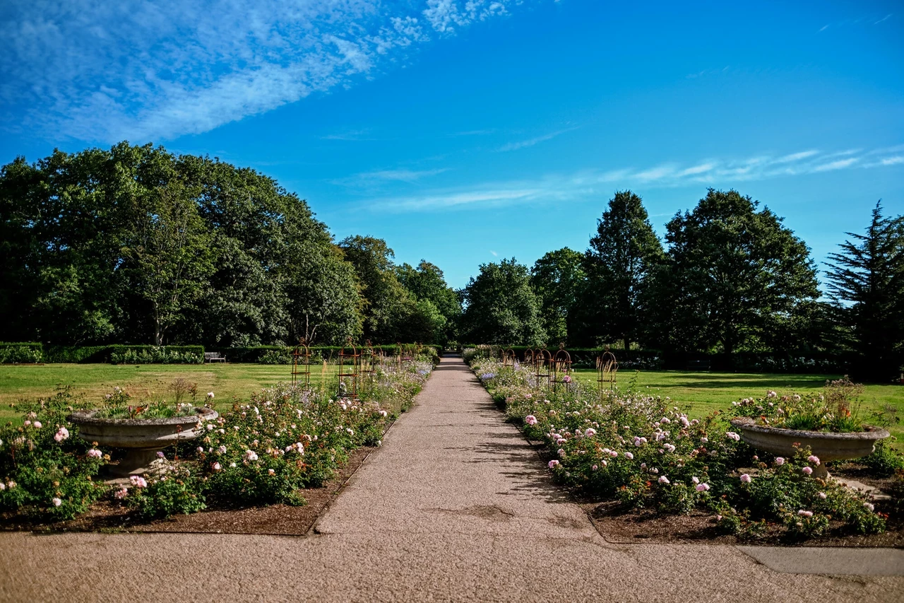 A wide pathway runs through a garden with flower beds of pink and white blossoms on either side. Stone planters and green lawns surround the path, while tall trees and a bright blue sky with light clouds. 