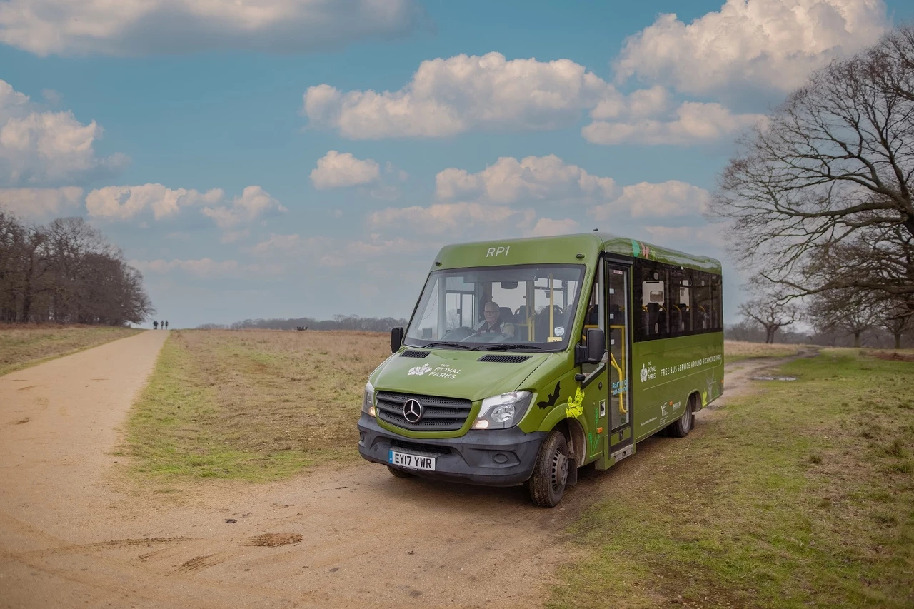 A green minibus on a dirt road in Richmond Park with a blue sky behind and white fluffy clouds